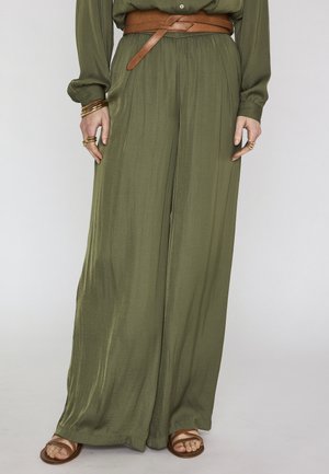 Donna che indossa pantaloni gamba larga verde oliva con una cintura in pelle marrone, braccialetti e anelli dorati, e sandali bassi marroni.