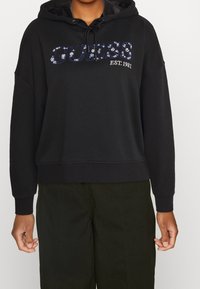 Sudadera negra con capucha ajustable, que presenta texto con lentejuelas "GUESS EST. 1981" en el frente. Corte holgado, tejido texturizado, dobladillo y puños acanalados.
