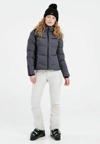 Schwarze, wattierte Jacke mit Reißverschluss, zwei Brusttaschen und schwarzer Strickmütze. Helle Hose und schwarze Skischuhe runden den Look ab.