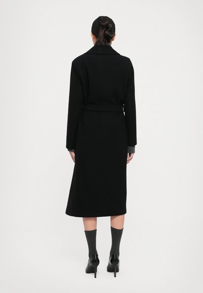 Woman standing with back to camera, wearing a long black belted coat, dark gray socks, and black high-heeled shoes against a plain background.