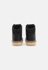 Zapatillas altas negras con un puño de tela texturizada, acentos de cuero liso y una suela de goma marrón. Se puede ver un detalle de costura reforzada.