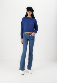 Suéter azul cropped com textura de malha espessa, combinando com jeans flare azul claro. Botas brancas de salto e um boina preta completam o visual.