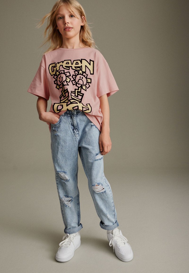 Rosa übergroßes T-Shirt mit cartoonhaften Blumen-Graphic, kombiniert mit hellblauen, ausgewaschenen Jeans und weißen Sneakers, vor einem neutralen Hintergrund.