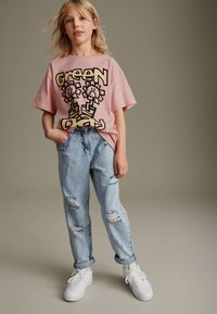 T-shirt oversize rosa con grafica di un fiore in stile cartone animato, abbinata a jeans chiari strappati e sneakers bianche, su uno sfondo neutro.