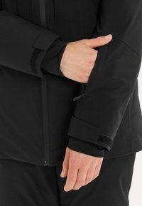 Svart vattentät jacka med en slät yta, med en dragkedjehals krage, kardborrearmar och en slim fit. Närbild av arm och hand.