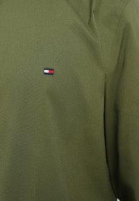 Olívazöld szövet kis téglalap alakú logóval, amely fehér, piros és tengerészkék színblokkokat tartalmaz.