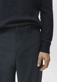 Pull en maille texturée bleu marine avec poignets côtelés, assorti à un pantalon sombre ajusté avec devant plat et poches latérales, main posée sur la hanche.