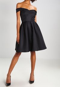 Black off-the-shoulder dress with a fitted bodice and pleated skirt. Made from smooth fabric, falls above the knees, complemented by high heels.