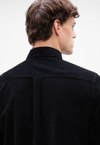 Mustan corduroy-paitapuseron korkea kaulus, jossa on taustayhdiste ja hienovarainen tekstuuri. Näkyvät ompeleet korostavat suunnittelua.