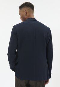 Tengeri blézer texturált anyagból, igényes szabással, klasszikus hajtókával és két hátsó szellőzővel. A hátsó nézet sima kialakítást mutat.