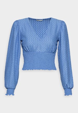 Blouse bleu clair avec un motif texturé, décolleté en V, manches longues bouffantes et taille smockée avec des bords en forme de vagues.