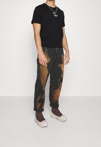 Musta puuvillainen T-paita, jossa valkoinen logo rinnassa, yhdistettynä mustiin farkkuihin, joissa on oranssinvärisiä batikkikuvioita, ja ruskeisiin korkeavartisiin saappaisiin.