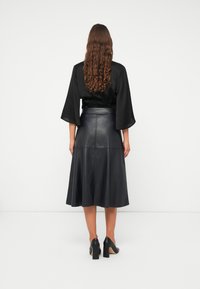 Blouse noire à manches longues avec une taille cintrée, associée à une jupe évasée en cuir noir. Le mannequin porte des chaussures à talons carrés noires.