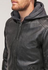 Giacca di pelle nera con zip frontale, caratterizzata da un cappuccio grigio scuro sottostante, texture liscia e dettagli di cucitura lungo le cuciture.