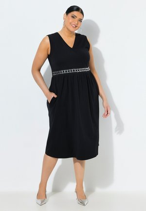 Smilende kvinde, der poserer i sort ærmeløs midi-kjole med pyntet taljebånd og sølv hæle foran ensfarvet, lyst baggrund.