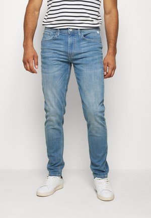 Mężczyzna noszący jasnoniebieskie obcisłe dżinsy, białe trampki i czarno-białą koszulkę w paski, stojący na jednolitym białym tle.