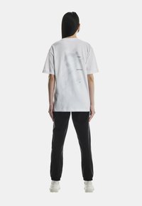 Kvinde med langt mørkt hår iført en hvid grafisk t-shirt, sorte bukser og hvide sneakers, stående med ryggen til foran en ensfarvet baggrund.
