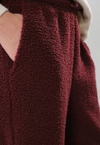 Burgunderfleece stoff, teksturert med en myk, hevet overflate. Nærbilde viser en sidelomme og detalj på linningen, som øker plaggets komfort.