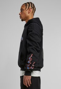 Sudadera negra de tejido suave con un bolsillo tipo canguro, diseños de olas coloridas en las mangas y un corte relajado.