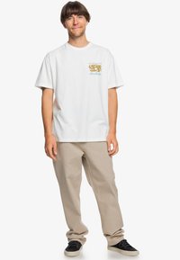 White short-sleeve t-shirt featuring a small colorful logo on the left chest. Paired with beige loose-fitting pants and black shoes.