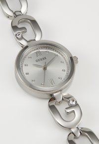 Silver Guess wristwatch with crystal hour markers, Roman numerals at 12 and 6, and a linked bracelet featuring crystal-embedded "G" logos.