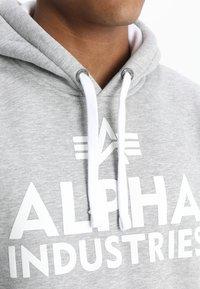 Sudadera gris hecha de una mezcla de algodón, con una gran impresión blanca de "ALPHA INDUSTRIES", cordones blancos y detalle del logo cerca del cuello.