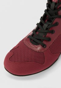 Scarpa sportiva rossa realizzata in mesh e suede, con lacci neri, punta tonda e dettagli testurizzati per traspirabilità e supporto.