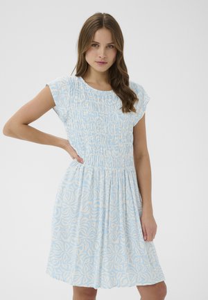 Kobieta ubrana w jasnoniebieską i białą sukienkę w krótkim rękawie z marszczonym gorsetem, stojąca z jedną ręką na biodrze na jednolitym tle.