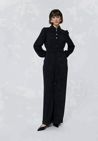 Γυναίκα στέκεται φορώντας μαύρη μπλούζα με μακριά μανίκια και κουμπιά από πέρλες, ψηλόμεσο παντελόνι με φαρδιά πόδια, μαύρη κορδέλα για τα μαλλιά και μαύρες μυτερές γόβες.