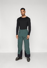 Calças de neve verdes com pernas inferiores pretas, múltiplos bolsos com zíper e fecho de botão. Combinadas com uma camiseta de manga longa preta ajustada.