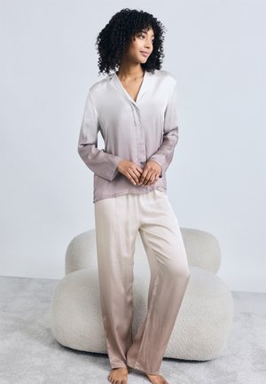 Γυναίκα με σγουρά μαλλιά που φοράει μεταξωτή πιτζάμα με διαβάθμιση χρωμάτων, στεκόμενη ξυπόλητη σε γκρι χαλί μπροστά από μια υφασμάτινη καρέκλα σαλονιού σε κρεμ απόχρωση.