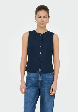 Kvinde med håret bundet tilbage iført en ærmeløs navyblå knap-top og blå jeans, stående mod en ensfarvet hvid baggrund.