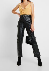 Black leather high-waisted cargo pants with zippers and pockets, paired with a mustard draped tank top. Model holds a black handbag.