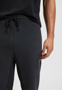 Dunkelgraue sportliche Jogginghosen mit einem Kordelzugbund, seitlichen Nähten und einem kleinen Markenpatch am rechten Oberschenkel. Glattes, dehnbares Material.