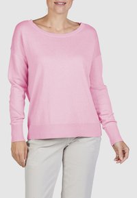 Vrouw in een losvallende roze trui met geribbelde mouwen en onderrand, gecombineerd met lichtgrijze versleten broek tegen een effen achtergrond.