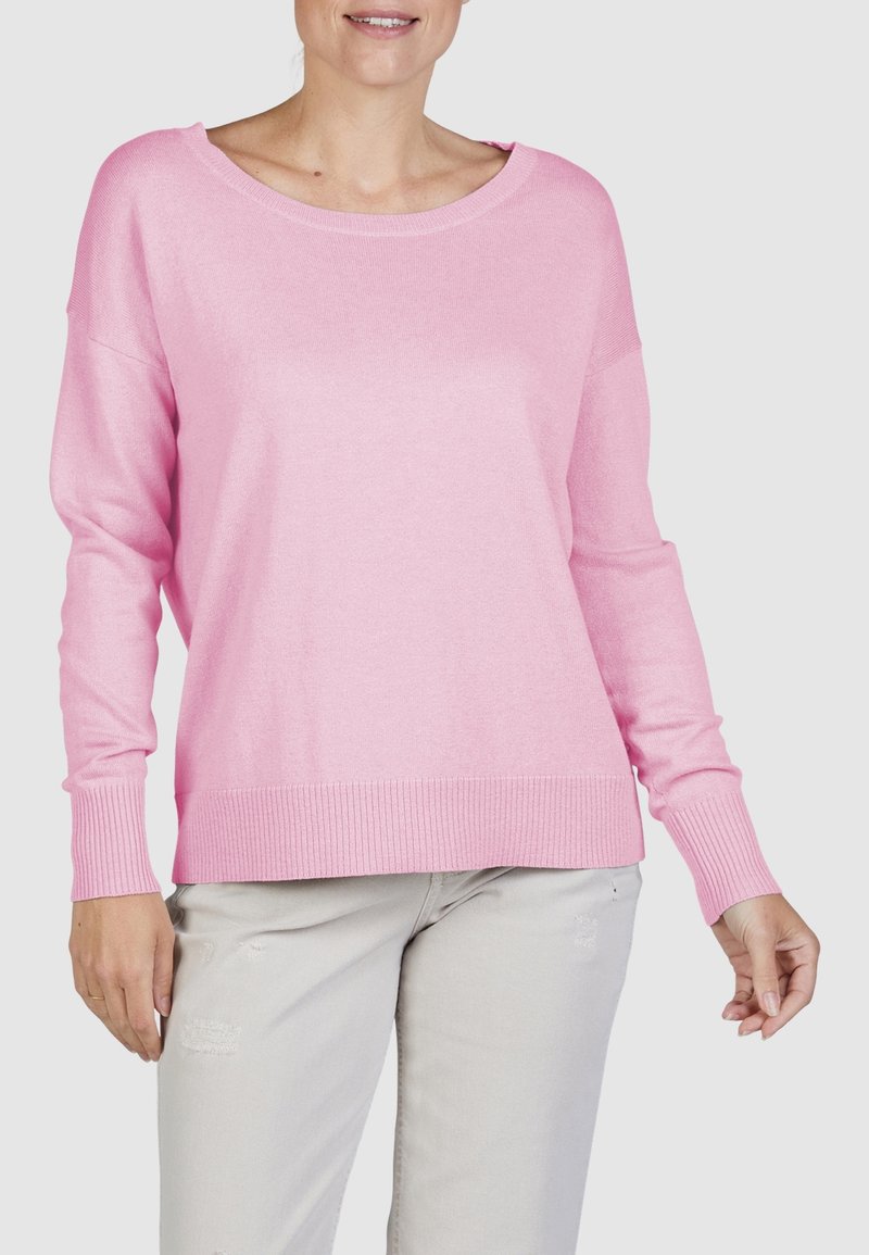 Vrouw in een losvallende roze trui met geribbelde mouwen en onderrand, gecombineerd met lichtgrijze versleten broek tegen een effen achtergrond.