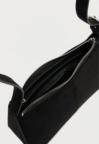 Geantă dreptunghiulară din piele întoarsă neagră, cu fermoar argintiu parțial deschis, care dezvăluie buzunare interioare și o curea scurtă cu cataramă.