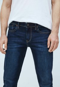 Mörkblå denimjeans med slim fit, som har en knäppning med knappar, fem fickor och kontrasterande sömnadsdetaljer.