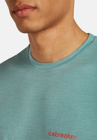 Teal kortærmet skjorte med rund halsudskæring. Har et rødt "icebreaker" logo på forsiden. Stoffet fremstår blødt og let tekstureret.