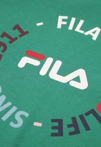 Camiseta de algodón verde con un gran logo blanco de "FILA", detalles en rojo y un patrón de texto circular en varios colores, sobre una textura suave.