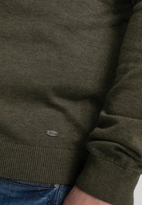 Olive green pullover sweater made of soft knit fabric; features ribbed cuffs and hem, along with a small metallic brand tag stitched on the side.