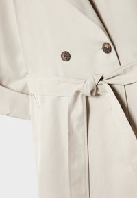 Gros plan d'un trench-coat beige montrant deux boutons foncés, une ceinture en tissu nouée et une partie d'une poche sur un tissu lisse.