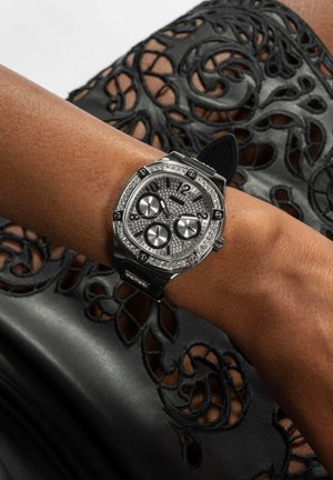 Czarny zegarek z srebrną ramką, trzema tarczami chronografu i diamentowymi akcentami, noszony na opalonej dłoni na tle strukturalnej czarnej koronki.