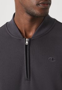 Suéter gris oscuro con cuello alto, frente con cremallera y un logotipo sutil. Textura suave con detalles de tejido acanalado a lo largo del cuello y la cremallera.