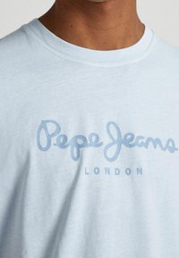 Světle modré tričko s krátkým rukávem vyrobené z měkké tkaniny, s stylizovaným logem "Pepe Jeans" tmavší modré barvy a nápisem "LONDON" uvedeným velkými písmeny pod ním.