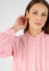 Blusa rosa con cuello con volantes y detalles de pliegues texturizados. El cierre de botones y las mangas fruncidas realzan el diseño. Tela suave y ligera.