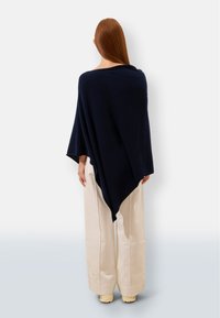 Granatowy poncho z miękkiego materiału o zwiewnym kroju, w kształcie trójkąta i z szerokimi rękawami, elegancko układające się na kremowych szerokich spodniach.