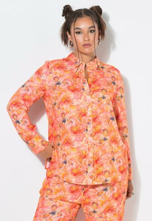 Frau mit zwei Dutts, die ein leuchtend orange-pinkes, geblümtes Hemd mit Knöpfen und passende Hosen vor weißem Hintergrund trägt.