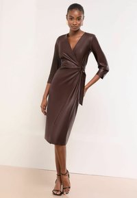 Rochie înfășurată din piele ecologică maro, cu decolteu în V, mâneci trei sferturi, detaliu cu legare pe lateral și tiv până la genunchi. Cizme negre cu barete la gleznă.