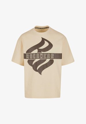 Vaaleanbeige puuvillainen t-paita, jossa on suuri ruskea grafiikkasuunnittelu ja "ROCAWEAR" -teksti edessä. Paita on varustettu pyöreällä kauluksella ja lyhyillä hihoilla.